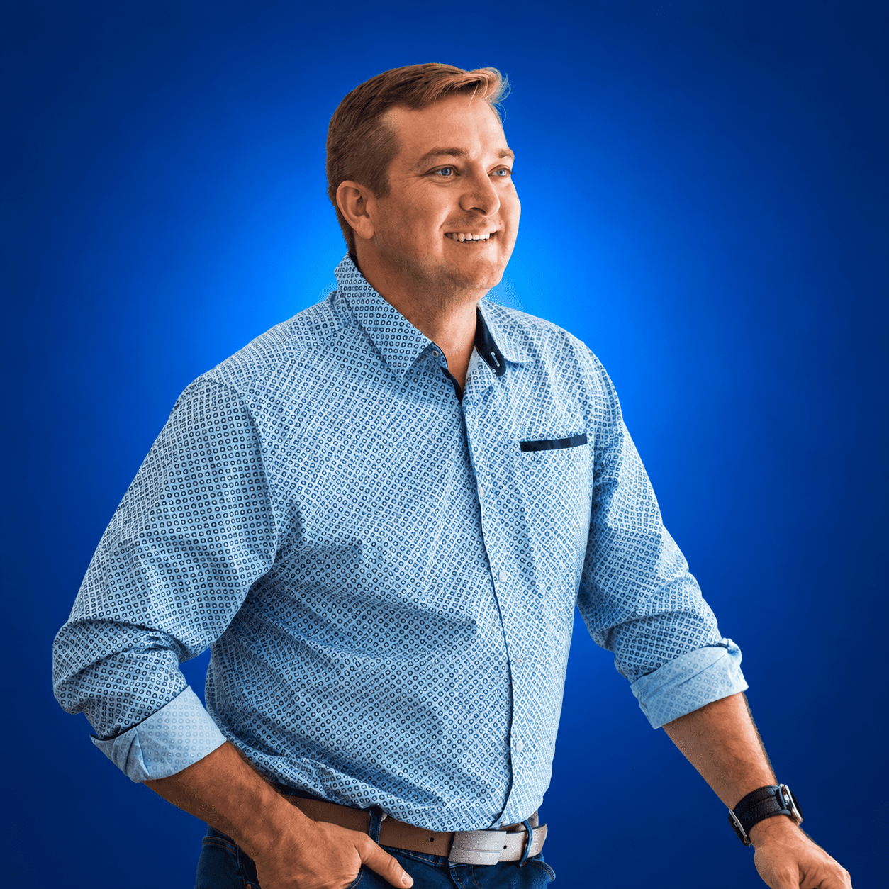 Mick Scotney smiling in a light blue patterned shirt against a blue background.